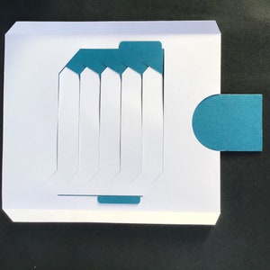 May include: A white card with a teal blue flap and a teal blue circle. The card has seven vertical slots cut out of it.