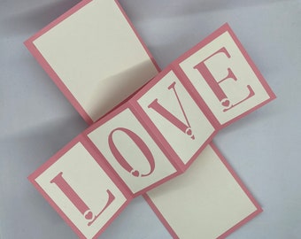 May include: A pink card with the word "LOVE" spelled out in white letters. Each letter is on a separate panel that folds out. Each letter has a small white heart in the centre.
