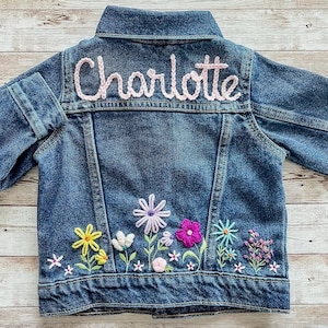 May include: A blue denim jacket with the name "Charlotte" embroidered in pink script across the back. The jacket features colorful floral embroidery along the bottom, including yellow, purple, and pink flowers.