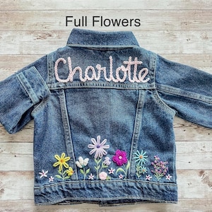 May include: A blue denim jacket with the name "Charlotte" embroidered in pink on the back. The jacket features colorful floral embroidery along the bottom, including yellow, white, purple, pink, and blue flowers. The text "Full Flowers" is at the top.