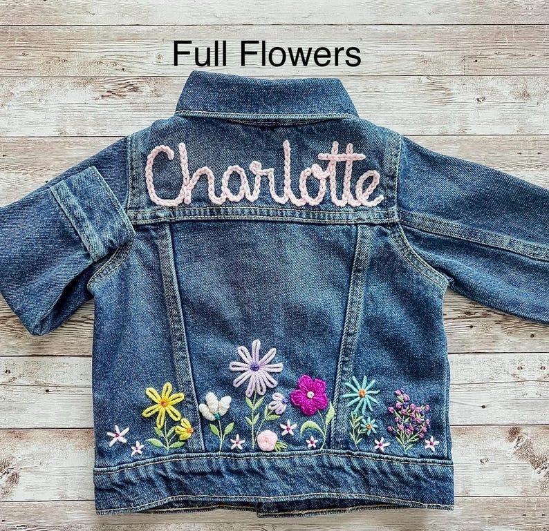 May include: A blue denim jacket with the name "Charlotte" embroidered in pink on the back. The jacket features colorful floral embroidery along the bottom, including yellow, white, purple, pink, and blue flowers. The text "Full Flowers" is at the top.