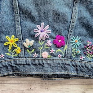 May include: A denim jacket with colorful embroidered flowers. The embroidery features yellow, white, pink, purple, and blue flowers with green stems and leaves. The jacket is a medium wash blue.
