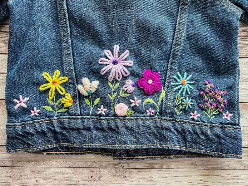 May include: A denim jacket with colorful embroidered flowers. The embroidery features yellow, white, pink, purple, and blue flowers with green stems and leaves. The jacket is a medium wash blue.