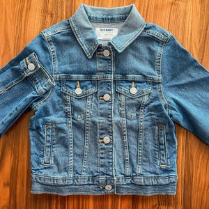 May include: A blue denim jacket with a classic collar, button closures, and two chest pockets. The jacket has long sleeves and the brand name "OLD NAVY" is visible on the label. The jacket is made of a sturdy denim material.
