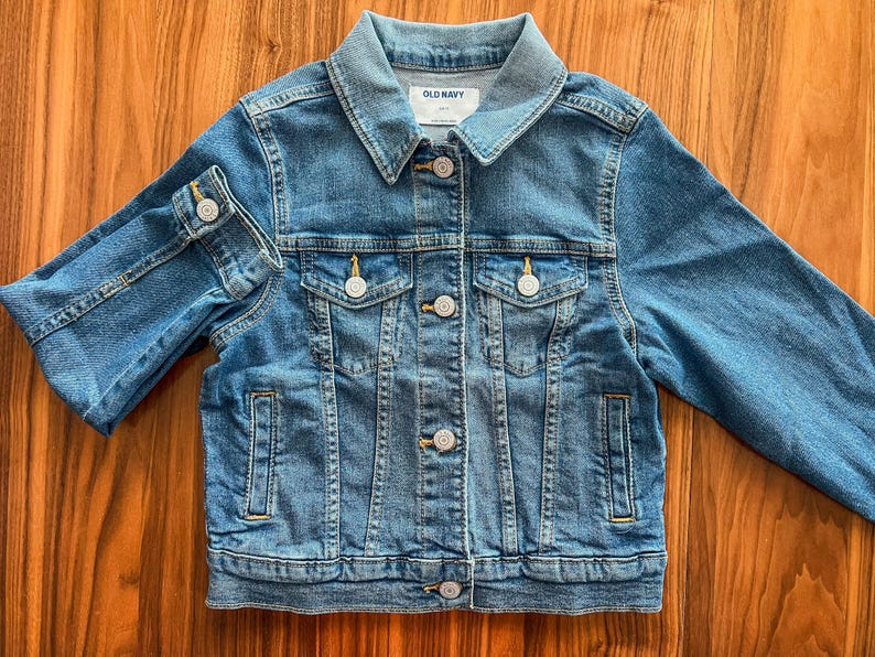 May include: A blue denim jacket with a classic collar, button closures, and two chest pockets. The jacket has long sleeves and the brand name "OLD NAVY" is visible on the label. The jacket is made of a sturdy denim material.