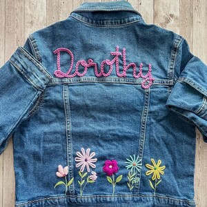May include: A blue denim jacket with the name "Dorothy" embroidered in pink script across the back. The jacket features colorful embroidered flowers along the bottom hem, including pink, purple, and yellow blossoms.