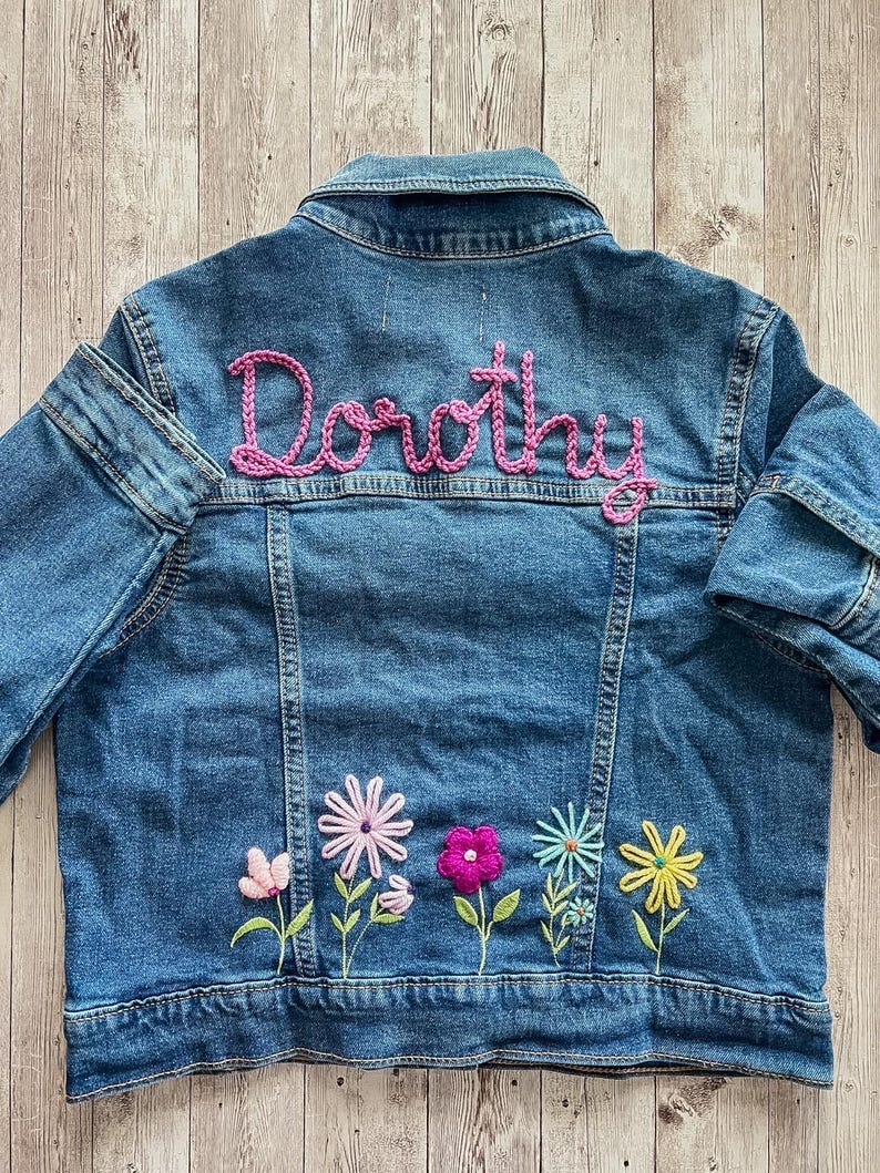 May include: A blue denim jacket with the name "Dorothy" embroidered in pink script across the back. The jacket features colorful embroidered flowers along the bottom hem, including pink, purple, and yellow blossoms.