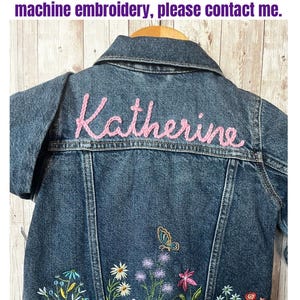 May include: A denim jacket with the name "Katherine" embroidered in pink thread on the back. The jacket features machine embroidery of flowers and a butterfly. The text on the image says "ONLY this design will be Hand-stitch Name with Machine Embroidery Flowers & Butterfly."