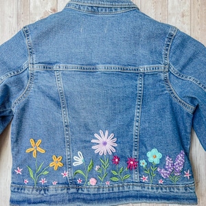 May include: A light blue denim jacket with embroidered flowers in yellow, pink, purple, and blue. The jacket is hanging on a wooden hanger. The floral embroidery is on the lower back of the jacket.