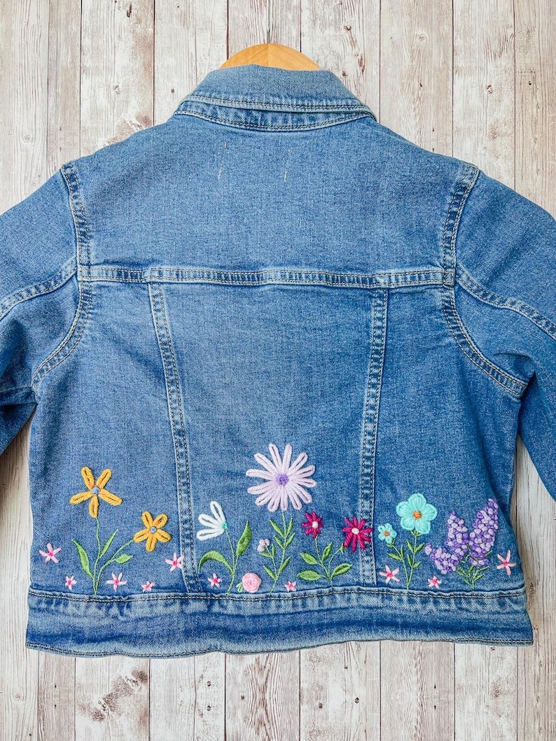 May include: A light blue denim jacket with embroidered flowers in yellow, pink, purple, and blue. The jacket is hanging on a wooden hanger. The floral embroidery is on the lower back of the jacket.