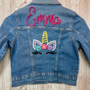 May include: A blue denim jacket with a unicorn design and the name "Emma" embroidered in pink. The unicorn has a yellow horn, pink and white face, and colorful flowers. The jacket is on a wooden hanger.