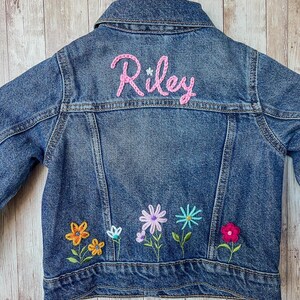 May include: A blue denim jacket with the name "Riley" embroidered in pink on the back. The jacket features colorful embroidered flowers along the bottom. The text "Simple Flowers" is at the top.
