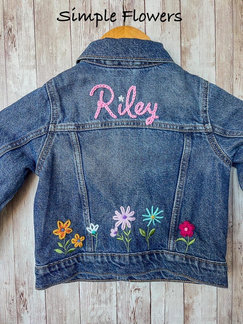 May include: A blue denim jacket with the name "Riley" embroidered in pink on the back. The jacket features colorful embroidered flowers along the bottom. The text "Simple Flowers" is at the top.