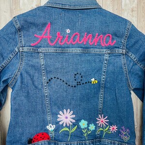 May include: Blue denim jacket with embroidered flowers, a ladybug, and a bee. The name "Arianna" is embroidered in pink on the back. The text "Flowers/Bee/Bug" is at the top.