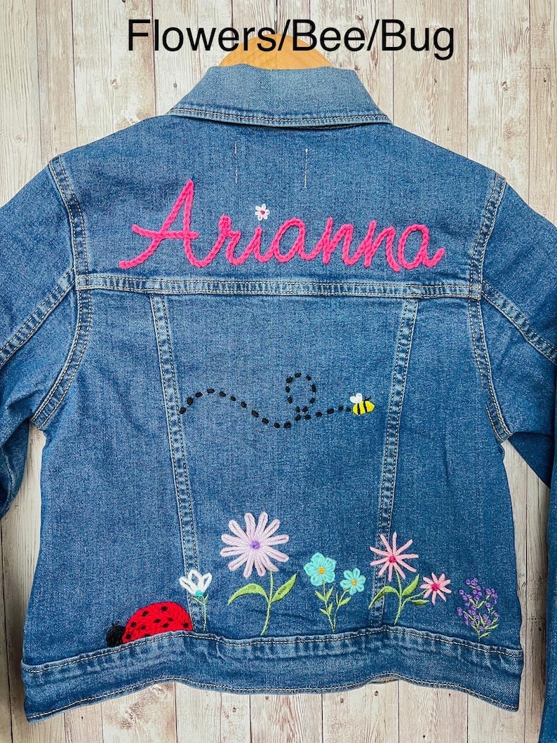 May include: Blue denim jacket with embroidered flowers, a ladybug, and a bee. The name "Arianna" is embroidered in pink on the back. The text "Flowers/Bee/Bug" is at the top.