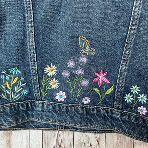 May include: Close-up of a dark blue denim jacket with colorful floral embroidery. The embroidery includes daisies, pink flowers, and a butterfly. The jacket has a classic design.