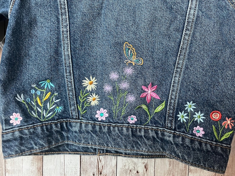 May include: Close-up of a dark blue denim jacket with colorful floral embroidery. The embroidery includes daisies, pink flowers, and a butterfly. The jacket has a classic design.