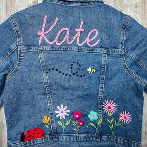 May include: A blue denim jacket with the name "Kate" embroidered in pink thread on the back. The jacket features a bee and a ladybug design, along with colorful embroidered flowers.