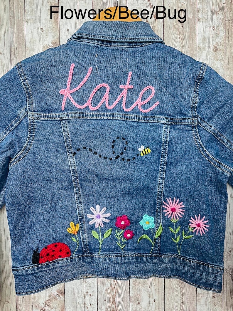May include: A blue denim jacket with the name "Kate" embroidered in pink thread on the back. The jacket features a bee and a ladybug design, along with colorful embroidered flowers.