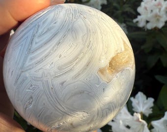 May include: A spherical stone with a polished surface, displaying a pattern of grey and white swirling bands. The stone has a smooth texture and a natural appearance. A section reveals a light brown, crystalline interior. The stone is held in a hand.