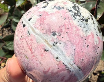 May include: A polished sphere of rhodochrosite, showcasing a blend of pink, white, and black patterns. The gemstone ball is held in a hand, with the focus on the spherical object. The surface has a smooth, reflective quality, highlighting the natural variations in color and texture.