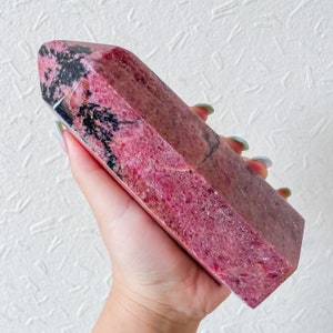 May include: A large, pink and black, polished,  faceted,  crystal point. The crystal is in the shape of a long, thin, rectangular prism.