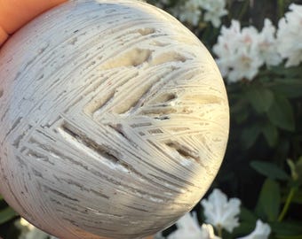 May include: A spherical, white and grey stone sphere with a unique, textured pattern of intersecting lines. The stone is held in a hand, with a blurred background of white flowers and green foliage.