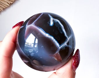 May include: A polished agate sphere with a dark purple and blue color pattern. The sphere is smooth and reflective.