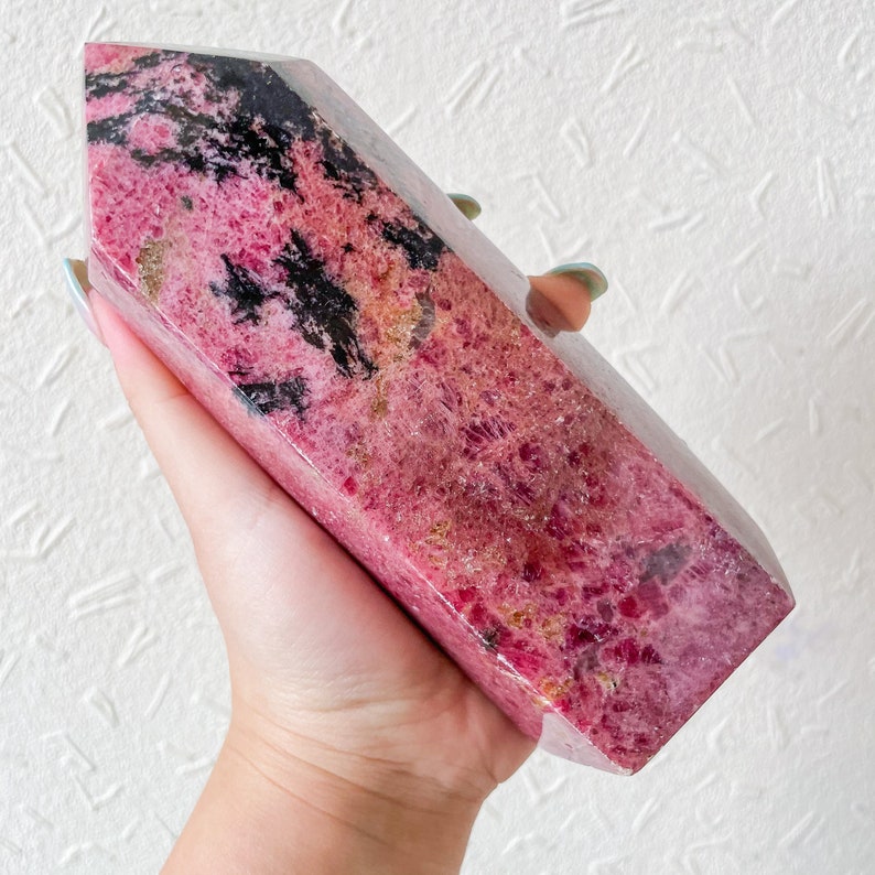 May include: A pink and black polished crystal point with a rough, natural texture. The crystal is a deep pink with black streaks and veins throughout. The crystal is held in a hand.