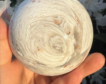 May include: A spherical stone with a swirling pattern of white and beige hues. The stone is held in a hand, highlighting its smooth, rounded form and natural variations. The surface of the stone has a distinctive, textured appearance.