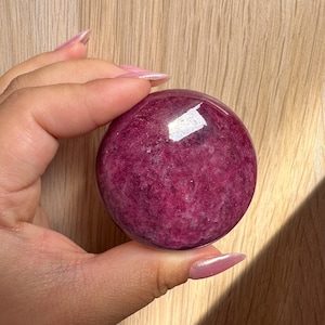 May include: A smooth, round, polished pink and purple gemstone. The stone has a swirling pattern and a glossy finish.