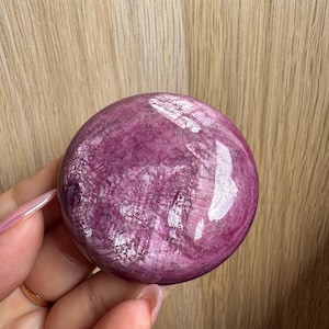 May include: A smooth, round, purple and white gemstone with a polished finish. The gemstone has a swirling pattern of white and purple.