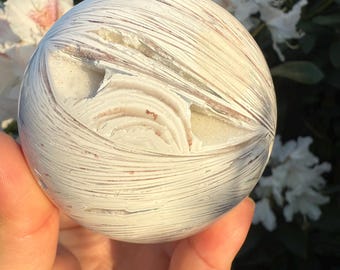 May include: A spherical, off-white stone with a distinctive pattern of fine, parallel lines and delicate reddish-brown shades. The stone's surface has a textured appearance, with areas of exposed, lighter-coloured material. The overall effect is reminiscent of natural formations.