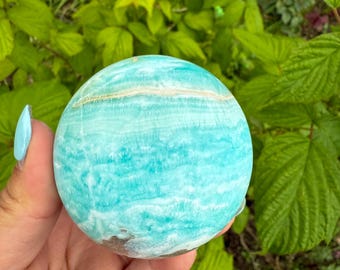 May include: A round, polished sphere of blue-green aragonite, with white and brown veining. The gemstone is held in a hand, with a background of green foliage. The sphere is approximately 3 inches in diameter, showcasing natural patterns and colors.