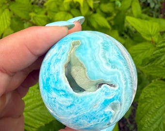 May include: A spherical, polished gemstone with a vibrant blue and white swirling pattern. The stone has a hollowed-out section revealing a lighter, beige interior. The gemstone is held in a hand against a backdrop of green foliage.