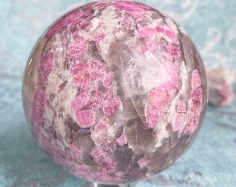 May include: A pink and grey crystal sphere with a swirling pattern of pink and grey inclusions. The sphere is smooth and polished.