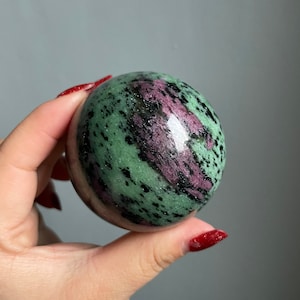 May include: A green, pink, and black sphere of polished zultanite gemstone. The sphere is smooth and has a high gloss finish.