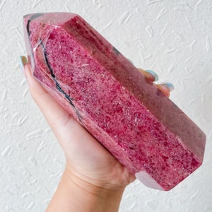 May include: A large, deep pink, polished, natural, raw, crystal point with black veins. The crystal is shaped like a long, thin pyramid.