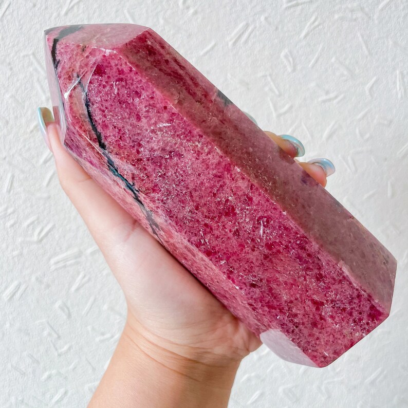 May include: A large, deep pink, polished, natural, raw, crystal point with black veins. The crystal is shaped like a long, thin pyramid.