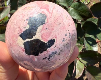 May include: A pink and black sphere, likely a decorative stone or crystal ball. The sphere has a smooth surface with a mix of pink and black patterns. The sphere is held in a hand, with green leaves in the background.