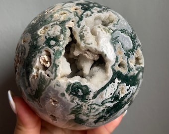 May include: A green and white agate sphere with a large, open cavity revealing a cluster of white crystals. The sphere has a smooth, polished surface and a natural, organic shape.