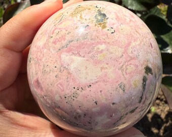 May include: A polished sphere of rhodochrosite, showcasing a blend of pink, white, and tan hues with dark gray speckles. The gemstone's surface is smooth, with a marbled appearance. The sphere is held in a hand.