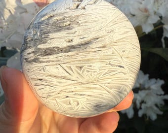 May include: A spherical stone with a distinctive pattern in shades of white and grey. The surface is marked with intricate lines and shapes, creating a textured appearance. The stone is held in a hand, with a backdrop of white flowers out of focus.