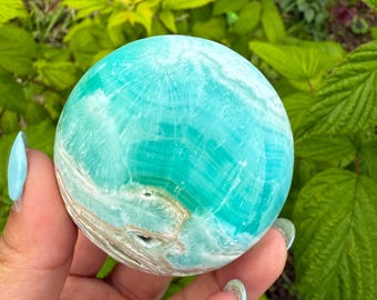 May include: A polished, spherical gemstone in shades of turquoise and white, with a swirling pattern. The stone is held in a hand against a backdrop of green foliage. The sphere appears to be approximately 3 inches in diameter.