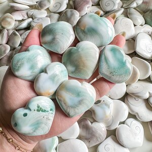May include: A collection of heart-shaped polished stones with swirling patterns in shades of white, green, and brown. The stones are arranged in a hand, showcasing their unique textures and colors.
