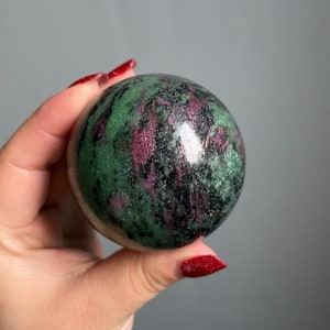 Puede incluir: Esfera de piedra preciosa verde, rosa y negra. La esfera está pulida y tiene un acabado brillante.