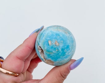 May include: A spherical, light blue gemstone with a marbled appearance. The stone has a few beige-colored inclusions and a small, irregular cavity. The sphere is held against a white background.