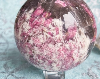 May include: A clear sphere with pink and white inclusions, resembling a geode, on a clear stand. The sphere is made of natural crystal.