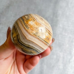 May include: A polished sphere of banded agate with brown, white, and gray stripes. The agate sphere is smooth and has a natural, organic look.