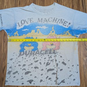 May include: White t-shirt with a colorful graphic design. The shirt features the text "LOVE MACHINE!" above a desert scene with cartoon figures and the Duracell logo. The shirt measures approximately 24 inches across.